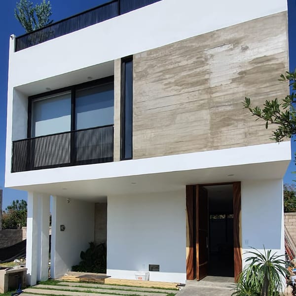 Fachada de dos niveles con muro de concreto texturizado, balcón, puerta de madera y jardín al frente.