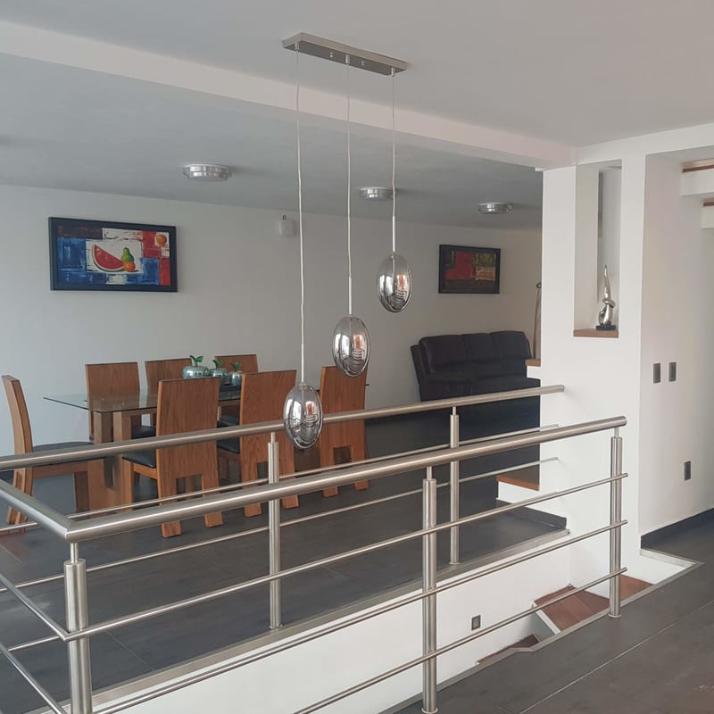 Interior de estancia con comedor de madera, barandal de acero y escalera al fondo.