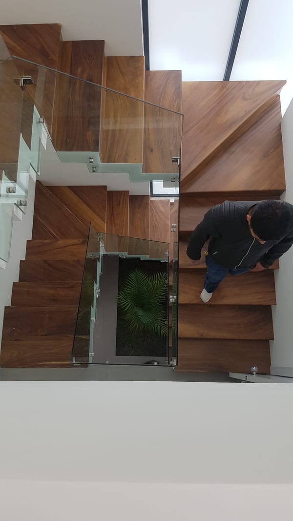 Escalera de madera vista desde arriba, con barandal de cristal y tragaluz.