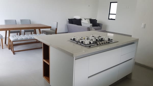 Estancia con comedor de madera, banca con cojines, sala y la isla de la cocina al fondo.