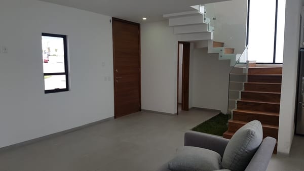 Interior con puerta de madera, escalera al fondo y sala junto al ventanal.