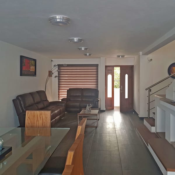 Sala con sillones de piel, comedor de cristal y la puerta principal al fondo.