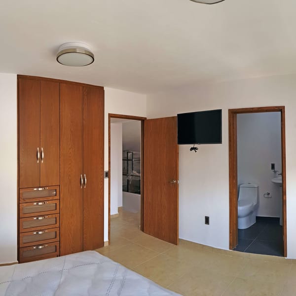 Recámara con clóset de madera empotrado, televisión en el muro y acceso al baño.